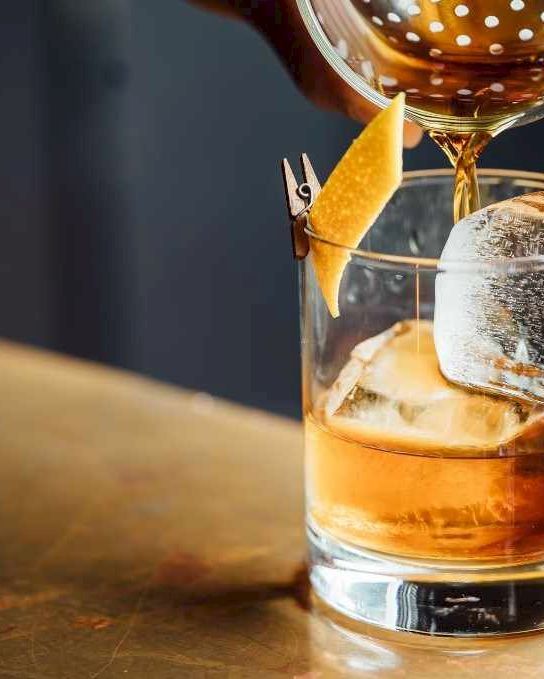 A whiskey on the rocks being poured into a glass with an orange twist garnish and a splash of garnish perched on the rim.